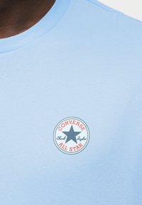 Ανοιχτό μπλε βαμβακερό t-shirt με στρογγυλό κέντημα που απεικονίζει ένα μαύρο αστέρι και κόκκινο κείμενο με τις λέξεις "Converse All Star" και "Chuck Taylor".