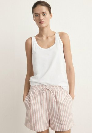 Ung kvinde med hænderne i lommerne iført hvid ærmeløs top og beige-stribede shorts med snor mod en enkel baggrund.