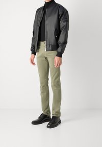 Man som bär en svart läderbombarjacka, svart tröja med hög krage, ljusgröna slim-fit byxor och svarta läderskor som står mot en vit bakgrund.