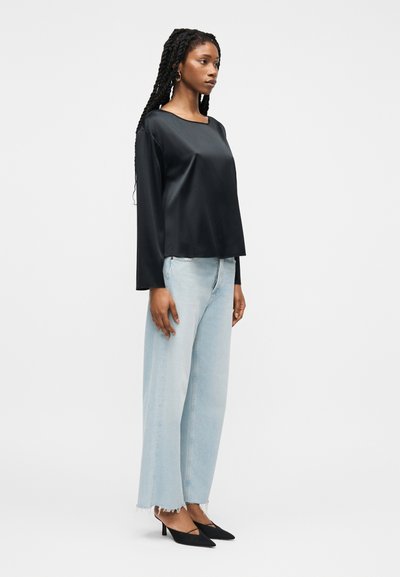 Γυναίκα με κοτσίδες στα μαλλιά, φορώντας μαύρο σατέν τοπ με μακριά μανίκια, ανοιχτό μπλε τζιν παντελόνι με φαρδιά μπατζάκια και γυμνές άκρες, και μαύρες γόβες με μυτερή μύτη, στέκεται πλαγίως.
