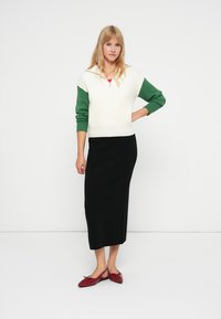 Pull blanc avec des manches vertes, col zippé, jupe longue noire et chaussures rouges. La tenue présente une coupe décontractée et une texture côtelée.