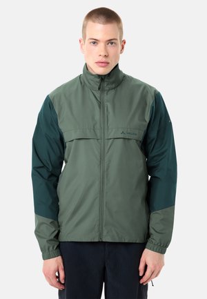 Junger Mann trägt eine grüne und dunkelgrüne Outdoor-Jacke mit Reißverschluss und hohem Kragen, steht vor einem einfarbigen hellen Hintergrund.