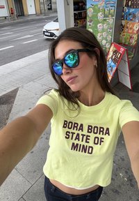 Žluté zkrácené tričko s nápisem "BORA BORA STATE OF MIND" v tmavě bordó barvě. Má krátké rukávy a volný střih. Černé sluneční brýle odrážejí okolí.