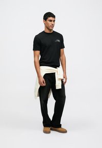 Czarny bawełniany T-shirt z małym białym logo, w połączeniu z luźnymi czarnymi dżinsami i beżowym swetrem związanym wokół talii. Brązowe obuwie.