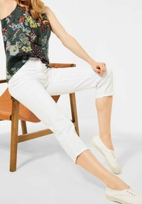 Floral-patterned dark green blouse with a twisted front, paired with white cropped pants and white slip-on shoes sitting on a wooden chair.