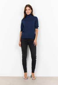 Pull en tricot bleu marine à col roulé, avec des manches courtes, associé à un jean noir slim et des sandales argentées à talons carrés. Texture lisse, design minimal.