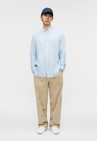 Man wearing blue striped button-up shirt, tan pants with dog prints, white sneakers, and navy blue baseball cap standing with hands in pockets.