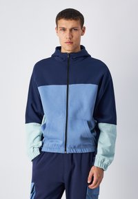 Felpa con cappuccio e zip in blu navy e azzurro chiaro con polsini a costine, caratterizzata da un design a colori divisi e tasche frontali. Texture morbida.