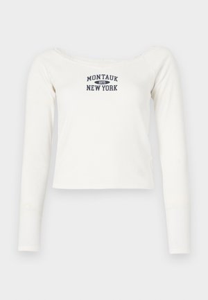 Top crop de manga longa branco, feito de um tecido suave, com um decote largo e uma impressão em azul marinho "Montauk 1975 New York" na parte da frente.