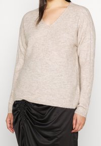 Beige, geribbelde gebreide trui met een V-hals, lange mouwen en een iets oversized pasvorm, gecombineerd met een zwarte gerimpelde rok.