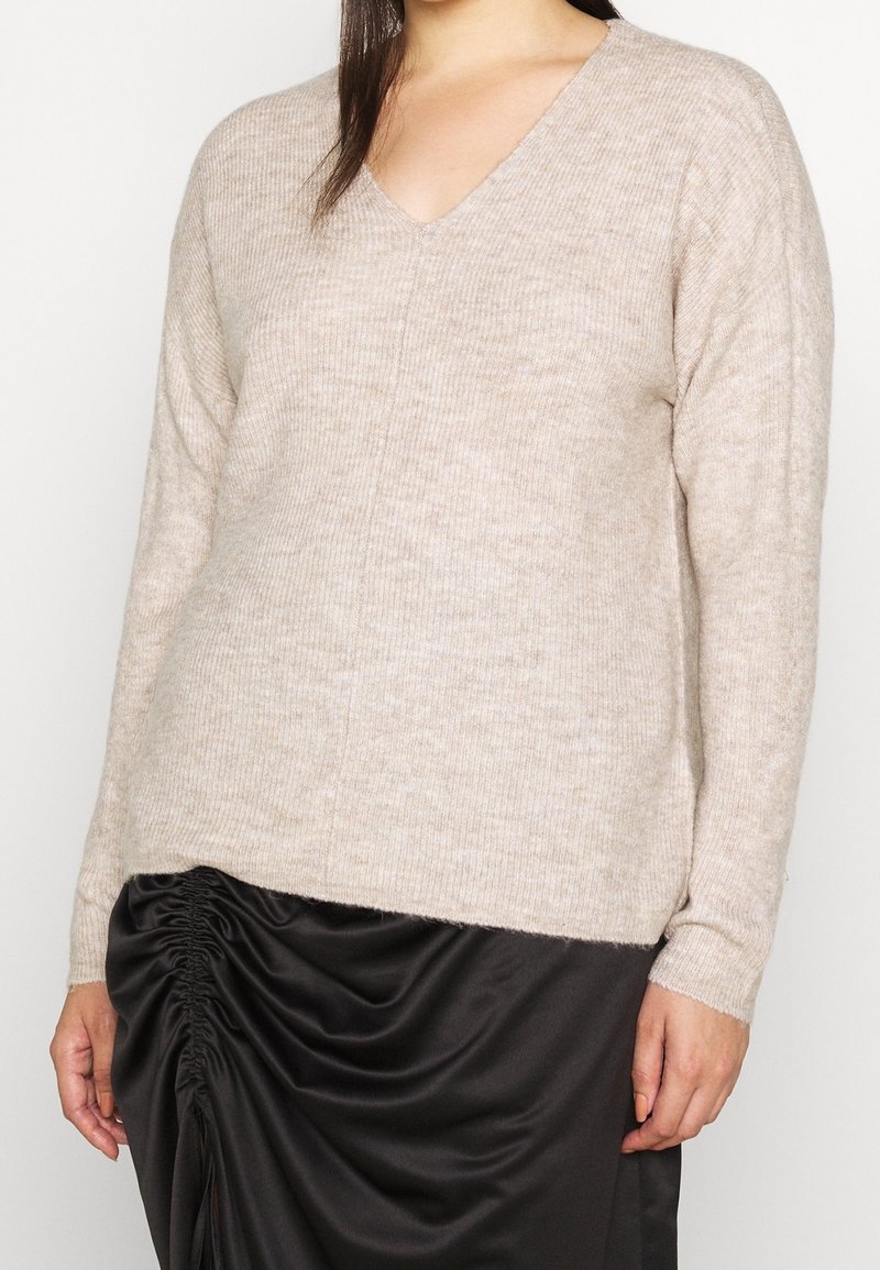 Beige, geribbelde gebreide trui met een V-hals, lange mouwen en een iets oversized pasvorm, gecombineerd met een zwarte gerimpelde rok.