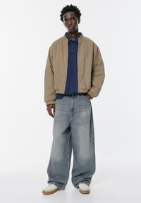 Giacca bomber beige sopra una polo blu navy, abbinata a jeans in denim chiaro a gamba larga e sneakers bianche con suola in gomma. Design semplice e vestibilità rilassata.