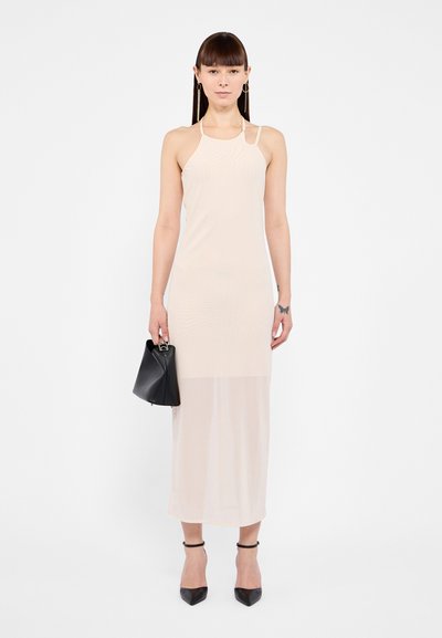 Beige sleeveless dress made of lightweight fabric, featuring a slightly sheer hem, worn with black pointed-toe heels and holding a small black bag.