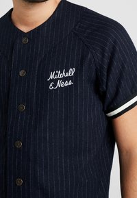 Marineblå skjorte med knapper, vertikale hvide nålestriber, rund krave, korte ærmer og broderet "Mitchell & Ness" logo på brystet.