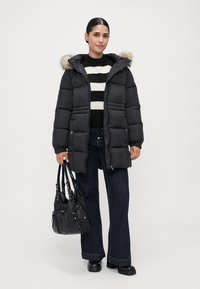 Mujer vestida con un abrigo de plumas negro con capucha de piel, suéter de rayas en blanco y negro, jeans oscuros, botas negras y sosteniendo un bolso negro.