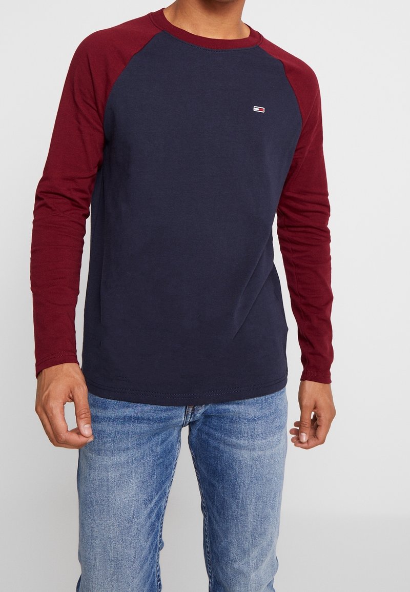 Pitkähihaisen t-paidan tummansininen vartalo ja burgundinpunaiset raglan-hihat. Rinnassa pieni logo. Valmistettu pehmeästä puuvillakankaasta.