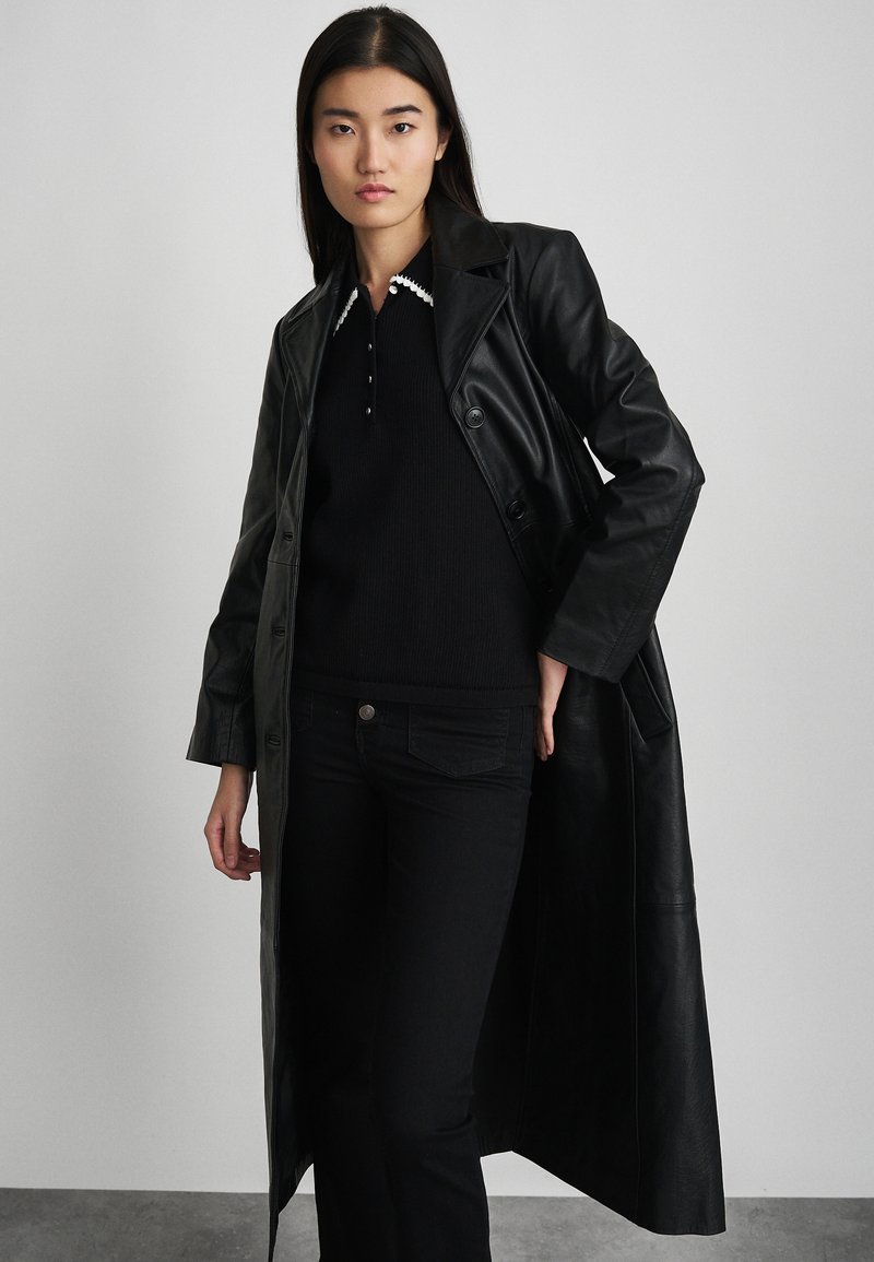 Cappotto trench in pelle nera con design doppiopetto, ampio colletto e tessuto texturizzato, abbinato a un maglione nero e jeans.