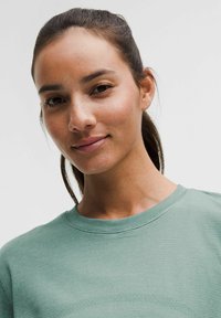 Sweatshirt verde deschis, cu guler rotund, având o textură subtilă. Prim-plan al feței unui model, punând în evidență nuanțele naturale ale pielii și machiajul minim.