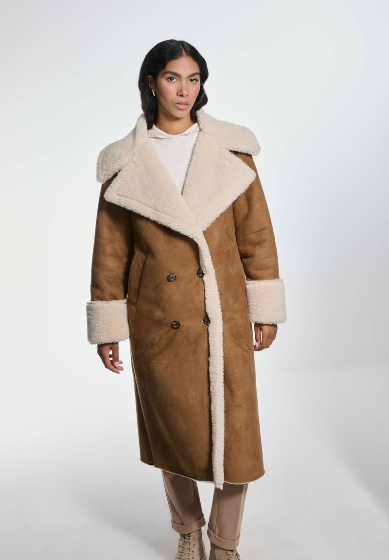 Brązowy płaszcz z faux zamszu z kremowym futrzanym kołnierzem i mankietami. Podwójnie zapinany design z dwoma przednimi kieszeniami i długim, strukturalnym kształtem.