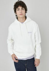 Haut à capuche en polaire blanche avec poche avant, cordons de serrage et texte violet clair. Porté par-dessus un t-shirt violet clair, associé à un jean foncé.