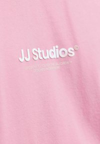 Camiseta de algodón rosa con el logo "JJ Studios" en texto blanco, con texto adicional blanco debajo. Textura suave y diseño casual.
