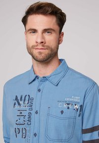 Camisa de botones azul claro con cuello, que presenta texto y diseños impresos, un único bolsillo en el pecho y acentos en color contrastante en las mangas.