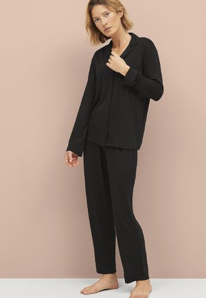Zwarte overhemd met lange mouwen en bijpassende broek van gladde stof, met een relaxte pasvorm en eenvoudig ontwerp, gedragen op blote voeten.