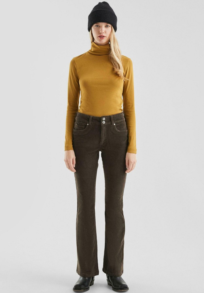 Un pull à col roulé moutarde associé à un pantalon en velours côtelé marron foncé évasé avec poches avant et fermeture à double bouton. Un bonnet en tricot noir est porté.
