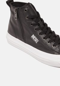 Mustat korkeavartiset tennarit, jotka on valmistettu sileästä nahasta. Niissä on valkoinen kumipohja, mustat nauhat, sivuvetoketju ja "DIESEL" -logotunnus.