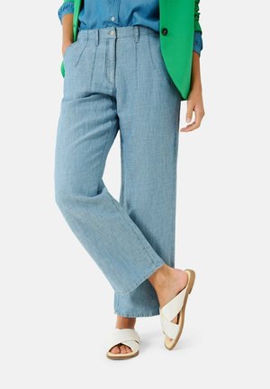 Femme portant un pantalon large bleu clair, une chemise bleue, un gilet vert et des sandales blanches croisées, debout avec une jambe croisée sur l’autre.