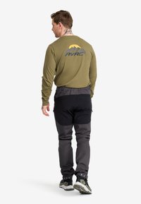 Man i en olivgrön långärmad skjorta med bergslogo och mörkgrå vandringsbyxor, går bortåt mot en slät vit bakgrund.