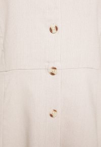 Chemise en tissu beige clair avec un tissage texturé, dotée d'une fermeture à boutons et de plusieurs boutons de couleur claire sur le devant.