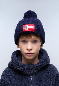 Cappello di lana blu navy con pompon e una patch rossa che presenta una croce bianca. Indossato con una giacca in pile blu navy con colletto alto.