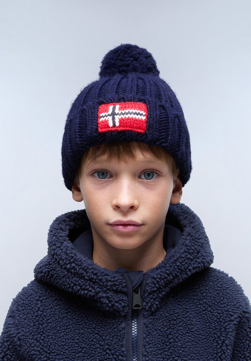 Cappello di lana blu navy con pompon e una patch rossa che presenta una croce bianca. Indossato con una giacca in pile blu navy con colletto alto.