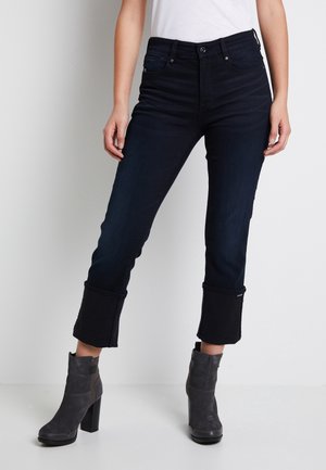 Jean en denim bleu foncé avec une taille haute, des jambes fuselées et des ourlets noirs. Associé à des bottines grises et une chemise blanche unie.