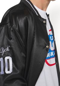 Person wearing a black satin varsity jacket with "Clyde 10" on the sleeve over a white graphic t-shirt with blue and red text.