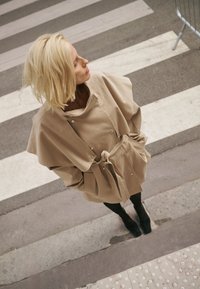 Szőke nő bézs színű ballonkabátban, kezeit zsebében tartva, egy gyalogátkelőhelyen áll oldalra nézve.