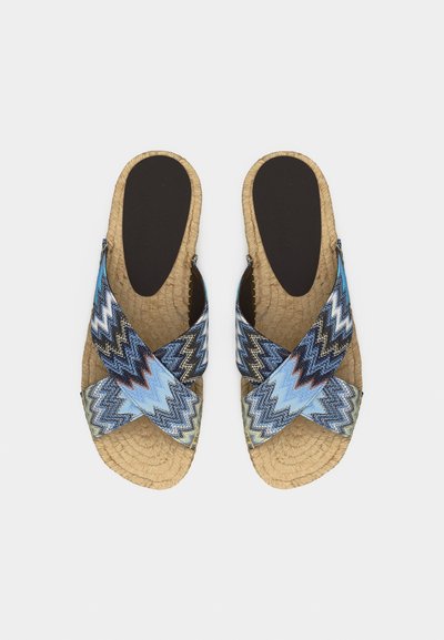 Ζευγάρι επίπεδων σανδαλιών με υφαντές μπεζ σόλες και σταυρωτούς υφασμάτινους ιμάντες με μπλε ζιγκ-ζαγκ σχέδια, απεικονιζόμενα από πάνω σε λευκό φόντο.