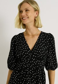 Vestido negro con lunares blancos, con escote en V, mangas abullonadas y cintura fruncida para una silueta fluida. Tejido texturizado.