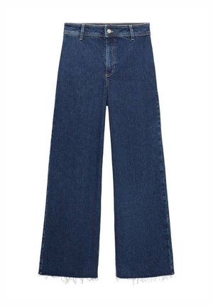 Person iført mørkeblå denimjeans og en sort jakke med den ene hånd placeret inde i baglommen på jeansene.