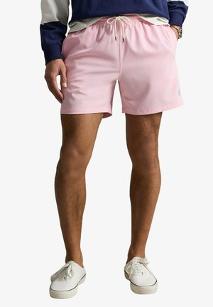 Hombre con pantalones cortos de cordón rosa claro, camiseta de manga larga azul marino y blanca, y zapatillas blancas, de pie con las manos en los bolsillos.