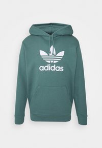 Huvtröja i teal-färgad tyg med en framficka i kängurustil, ribbade ärmslut och nederkant, samt en stor vit Adidas-logotyp med tre ränder.