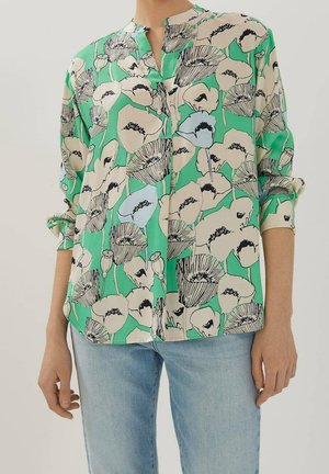 Nainen, joka pukeutuneena vihreään kukkakuvioiseen pitkähihaiseen paitaan, hihansuut käärittyinä, ja vaaleansinisiin farkkuihin, seisomassa yksiväristä taustaa vasten.