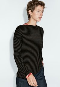 Giovane con capelli corti indossa un maglione a righe nere e grigie sopra una camicia a righe rosse e bianche, abbinato a pantaloni scuri, su sfondo bianco.