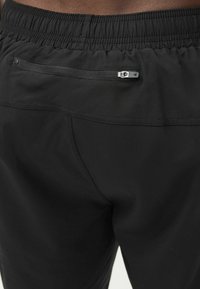 Svarta elastiska byxor med resår i midjan, en dragkedja på bakfickan och en triangulär logotyp på dragkedjens dragare.