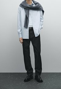 Camicia azzurro chiaro con bottoni e maniche lunghe, abbinata a jeans in denim scuro e un maglione di lana grigio scuro portato sulle spalle. Scarpe nere.