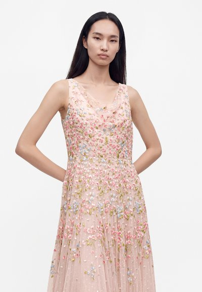 Femme aux longs cheveux foncés portant une robe sans manches rose pâle avec de petites broderies florales bleues, roses et vertes sur fond blanc.