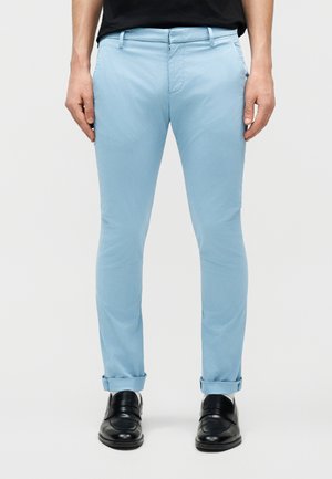 Άνδρας που φοράει στενά, ανοιχτό μπλε παντελόνια με γυρισμένα μανίκια και μαύρα επίσημα παπούτσια, στέκεται μπροστά σε απλό φόντο.