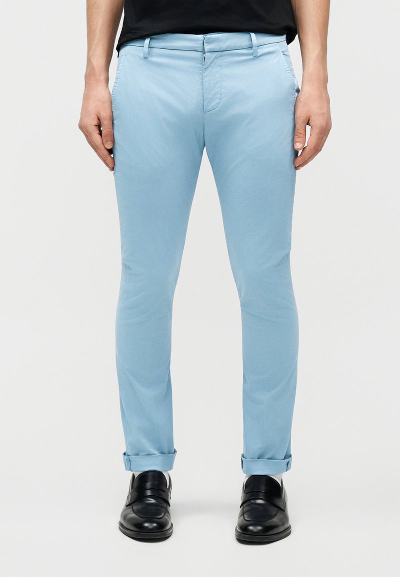 Άνδρας που φοράει στενά, ανοιχτό μπλε παντελόνια με γυρισμένα μανίκια και μαύρα επίσημα παπούτσια, στέκεται μπροστά σε απλό φόντο.