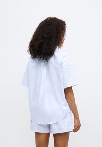 Ριγέ κοντομάνικο πουκάμισο σε ανοιχτό μπλε με κάθετες λευκές γραμμές, με στρογγυλεμένη τσάκα και άνετη εφαρμογή, συνδυασμένο με ταιριαστά σορτς.